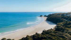 Aerial view of Byron Bay’s main beach with turquoise waves and lush headland under a clear blue sky