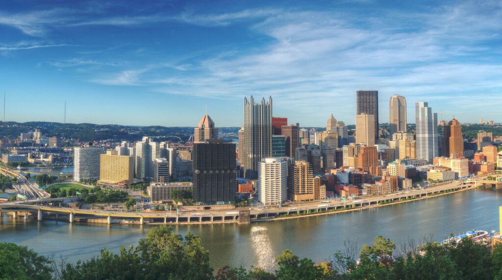 Panoramic Pittsburgh skyline with downtown skyscrapers and yellow bridges above the Three Rivers, framed by green hillsides in Pennsylvania.